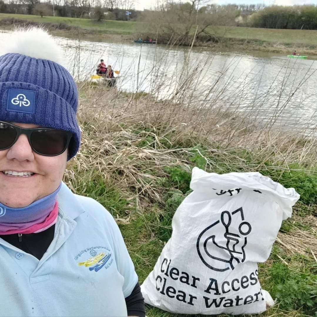 A woman with sunglasses on and a bobble hat stands by a river holding a Clear Access Clear Water bag