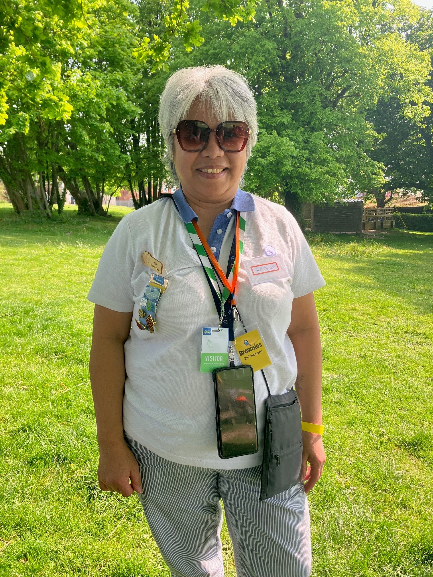 A woman with short grey hair smiles at the camera. She is standing in a green field, wears sunglasses and lanyards against a Girlguiding top and animal print trousers