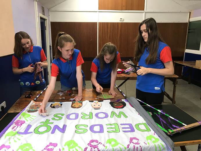 Four Guides painting a banner featuring the slogan "Deeds not words" in women's suffrage colours of green and purple