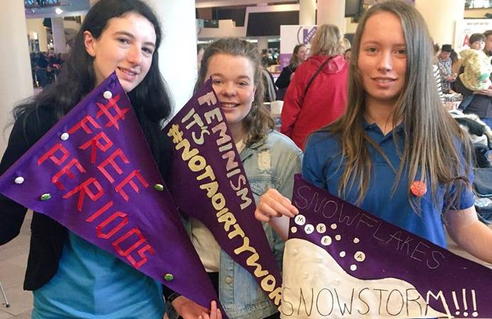 Three young women holding handmade flags - one flag says "free periods", another "Feminism is not a dirty word" and another "snowflakes make a snowstorm"