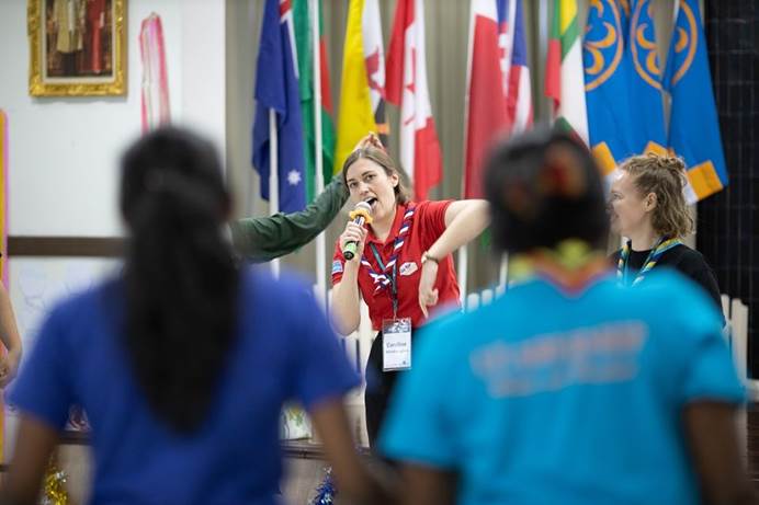 GOLD coordinator Caroline Brown wears red polo shirt and neckerchief and is holding a mic, surrounded by other GOLD project members and against a backdrop of flags