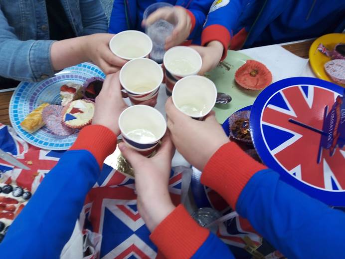 6 Guides' hands cheers cups over a buffet. You can see napkins, a cake stand, plates of food and lots of Union Flags