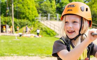 A smiling blonde girl in a Brownie uniform, harness and PGL helmet, gets ready to climb a tall pole.