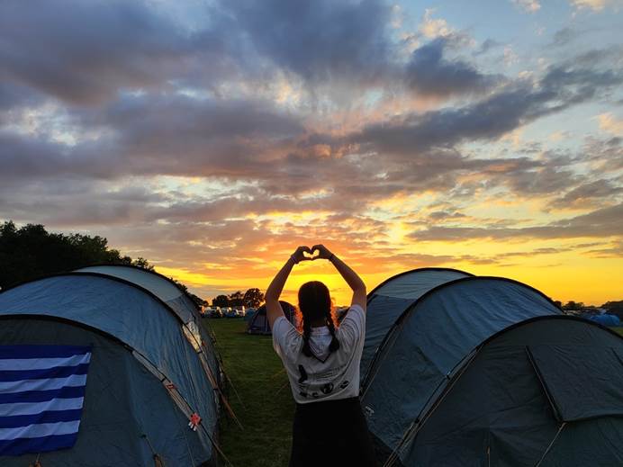A Greek team member watches the sunset whilst holding her hands up in a heart shape in a campsite