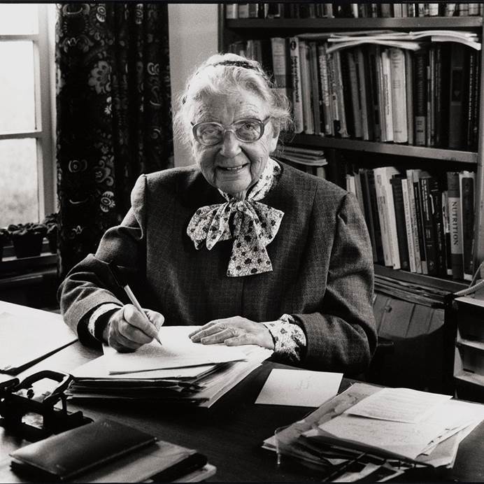 A black and white image of Elsie Widdowson sitting at her desk with a bookcase behind her