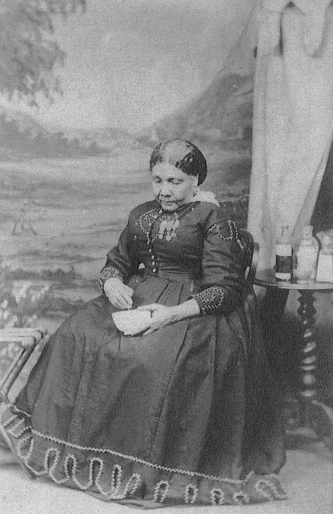 A black and white photograph of Mary Seacole who is looking down and mixing something in a bowl. Next to her is a table with glass bottles on it.