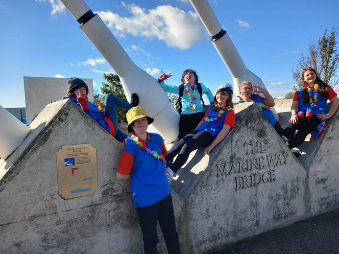 Guides from 1st Skelmersale Guides sit on the triangles of The Marine Way Bridge sign in Southport, wearing hats and garlands and smiling and pulling faces