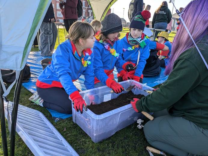 Three guides in uniform, hats and garlands look into a box of dirt and a bone they have dug out of it.
