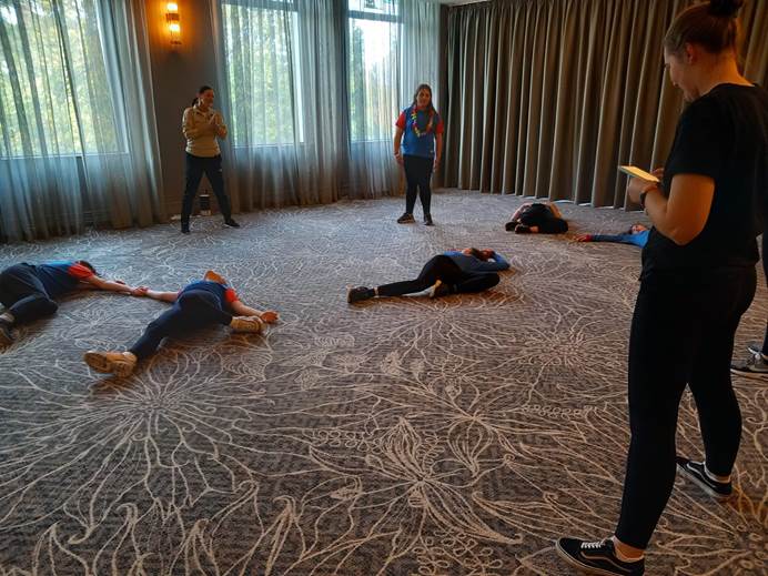 In a room a tutor leads a tai chi workshop with 5 guides on the floor in poses while one stands up.