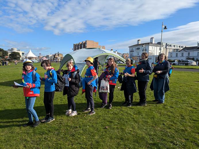 A line of guides wearing coats and hats and carrying clipboards stand in a park with the leader wearing a captain's hat ready to leave for the next challenge.