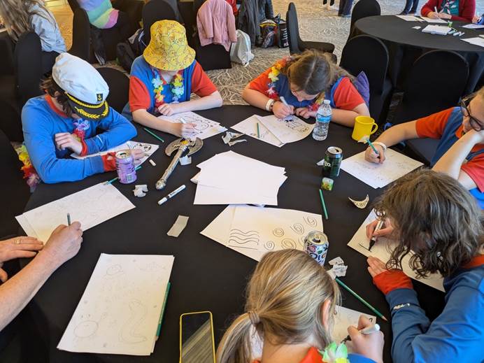 6 guides sit around a circular table indoors drawing swirls and sea creatures using marker pens on white A4 paper.