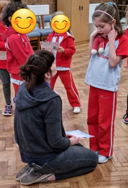 Sophie, who's white, has brown hair in a bun, is wearing a grey hoodie and grey jeans, kneels on the floor in front of Agnes, holding a Rainbow promise certificate. Agnes, who's white and has light brown hair in a ponytail and a pink hairband and is wearing Rainbows uniform, looks down at Sophie saying her promise, with her fingers in her ears and holding an Olivia doll