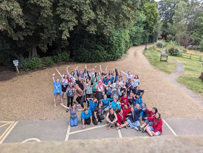 Members of Girlguiding's advocacy group Amplify join GOLD teams in a big group and wave at the camera in a space outside.