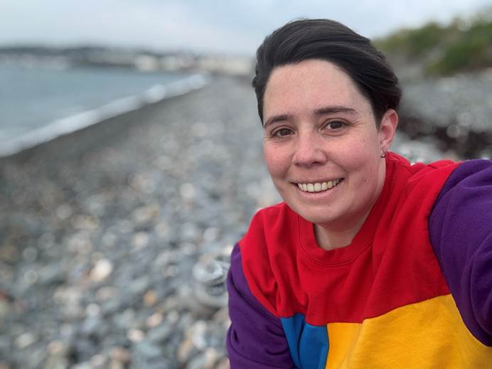 A woman with short brown hair and a yellow, red and blue jumper sits on a stony beach smiling at the camera