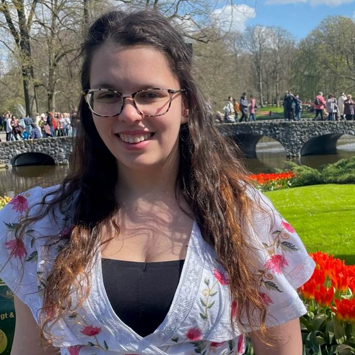 Bee stood in a grassy outside area. She's white, has long, wavy brown hair, glasses and is wearing a white top. She's smiling at the camera