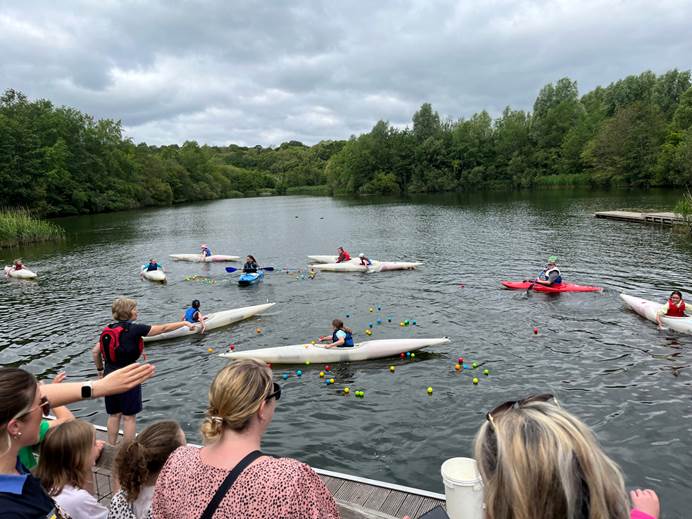 10 canoes are on the water with Brownies playing duck race, watched by their leaders and parents from the shore