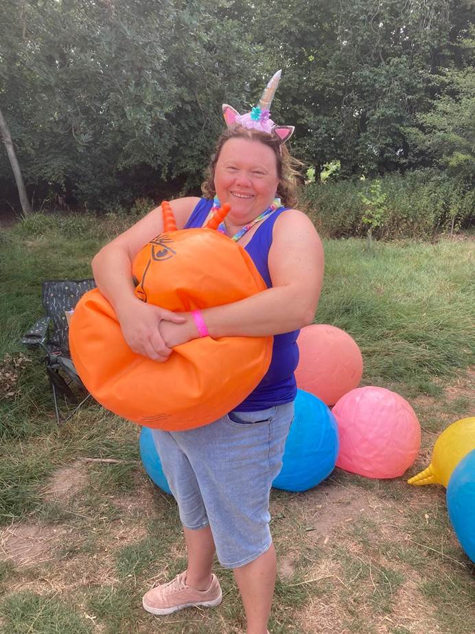 Caroline stands alone smiling at the camera. They're holding a deflated space hopper and wearing a unicorn headband. Behind them are more deflated hoppers, grass and trees