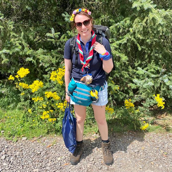 Sally Kettle, deputy chief guide, wearing Girlguiding uniform and a blue bum bag. Sally is a white mid height woman and is wearing a flower crown and black sunglasses. She's smiling at the camera and standing outdoors in a sunny field