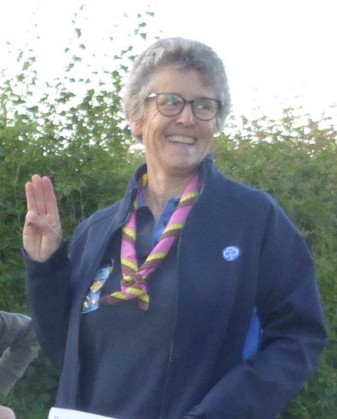 A woman with grey hair wearing a leader jacket and necker smiles and makes the Brownie promise sign