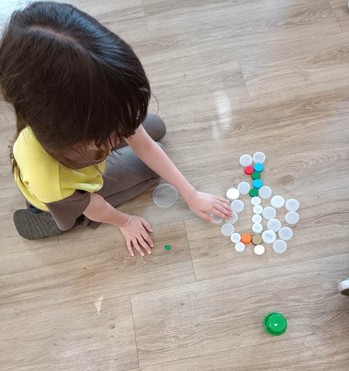 A Brownie sitting on the floor making a sea creature shape from plastic bottle tops