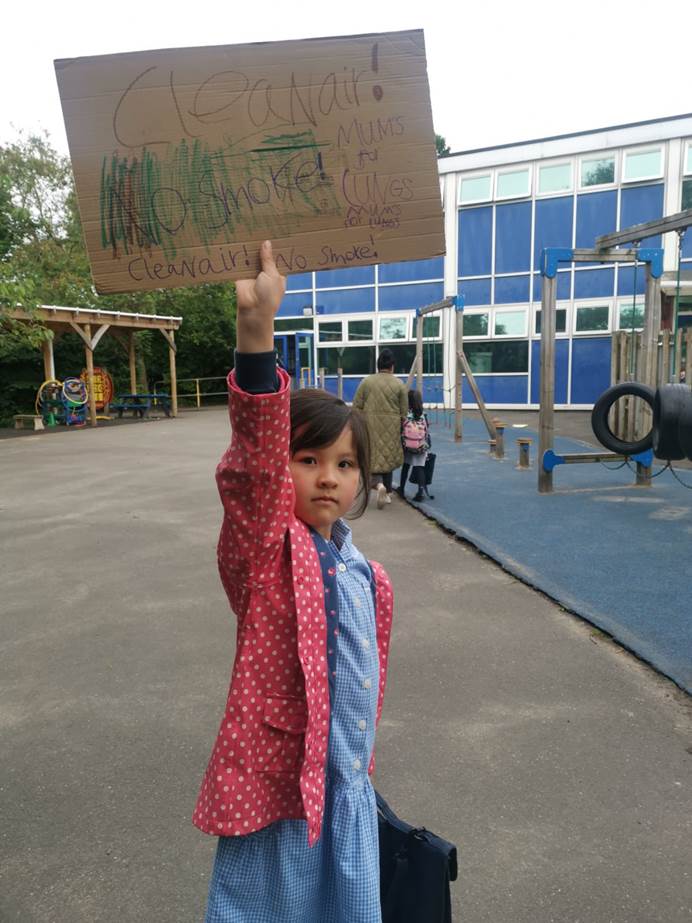 A Rainbow standing outside her school holding a home-made sign which says 'Clean air! No smoke!'