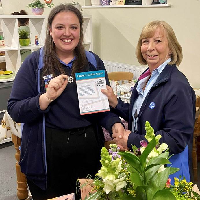 Georiga is on the left and another member is on the right. They're stood inside and there are lots of decorations. Georgia is shaking hands with the member, holding a badge and certificate. They're both white and wearing uniform and smiling. Georgia has long, brown hair