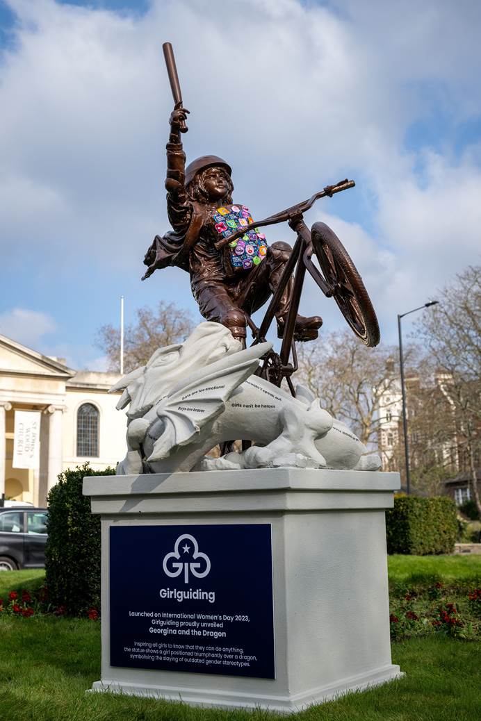 A bronze statue of a girl riding a BMX. She's doing a wheelie, carrying a backpack with Girlguiding badges on, and holding a baseball bat above her head. The BMX is on a white sculpted dragon, with outdated stereotypes written on it. The sculpture is on a plinth with the refreshed Girlguiding logo on it