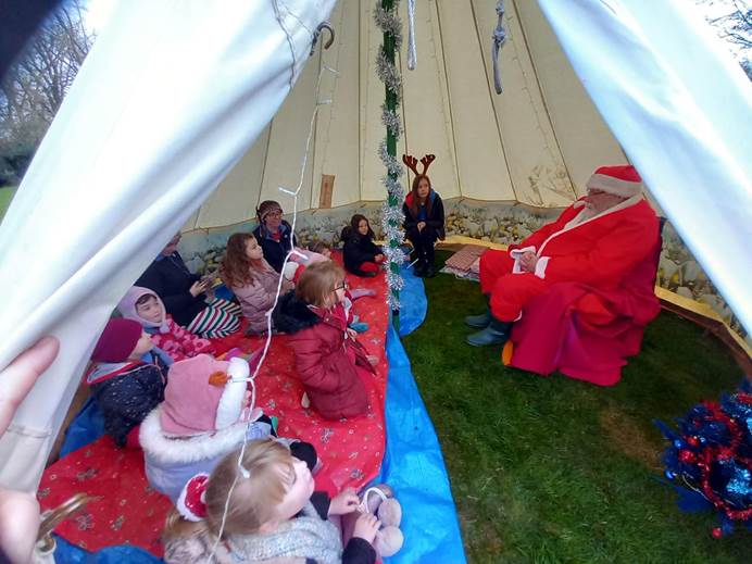 A group of Rainbows and their leaders in coats sit in a tent on blankets and meet Santa, a man with white hair in a red suit