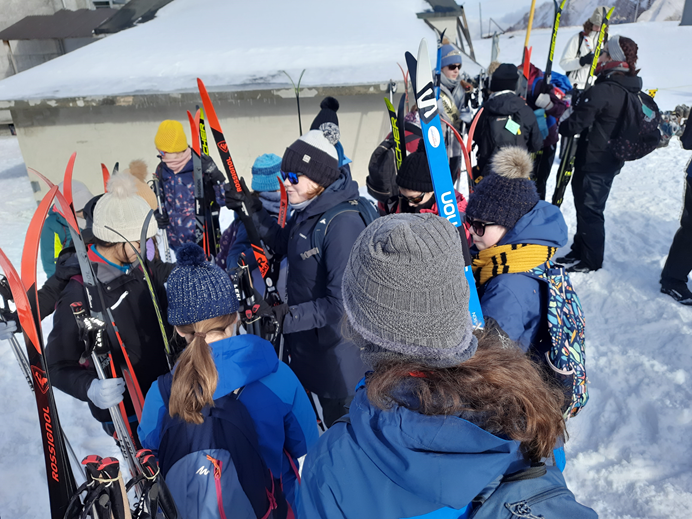 A group of young and older women stand in the snow in Switzerland, wearing ski wear, skis and bobble hats getting ready to ski