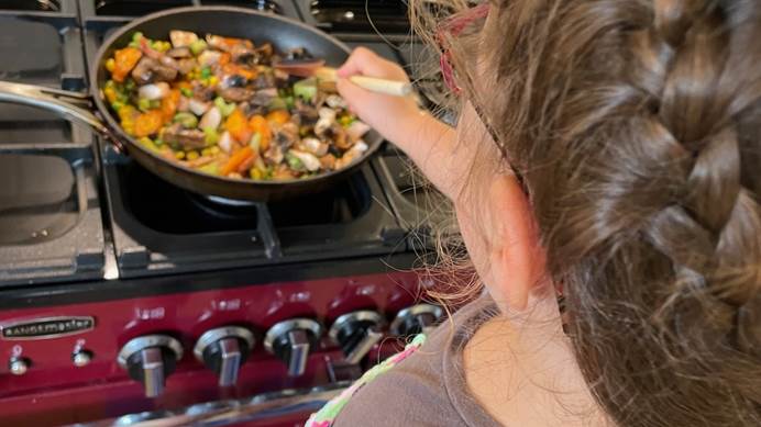 A young girl with brown hair, back towards the camera. Stood in front of a stove and stirring a frying pan full of vegetables. 
