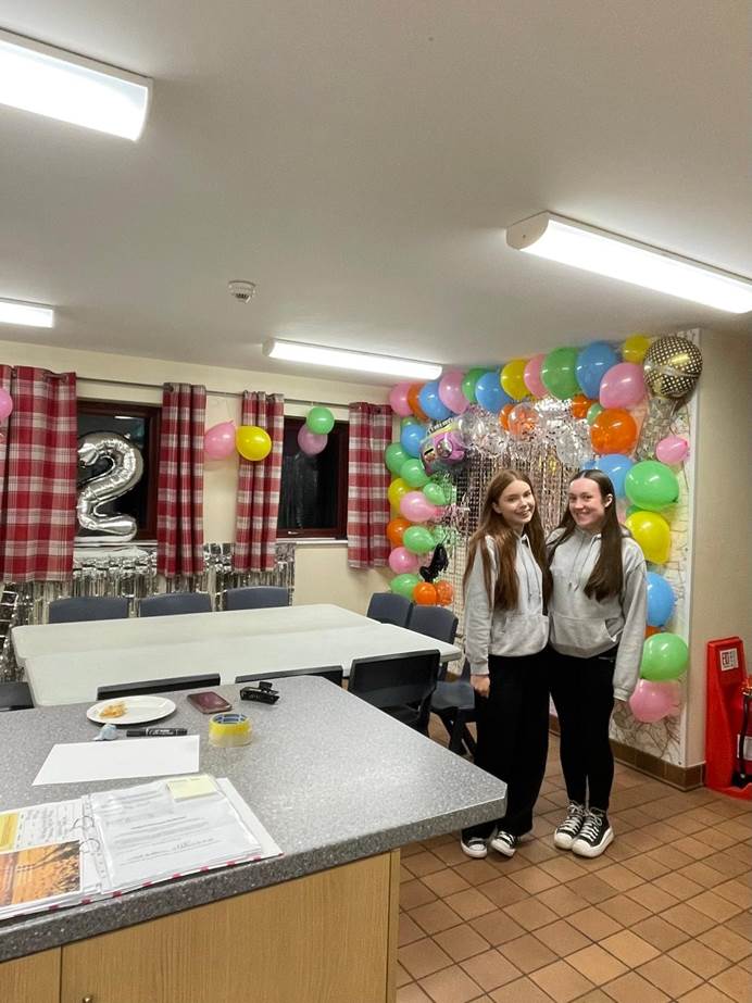 Two brown haired girls in grey hoodies and black leggings stand together in a room decorated for a party and smile at the camera