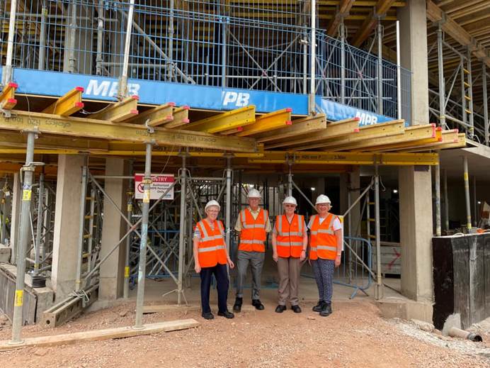 A woman and three men stand in orange high vis jackets and white hard hats, looking at the camera. Surrounding them is building work