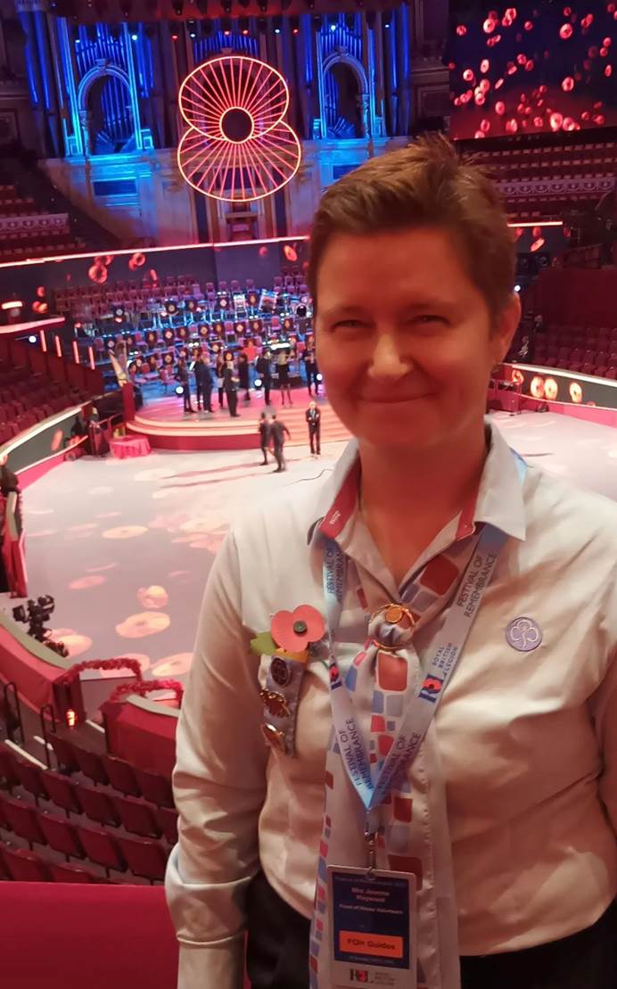 Jo stands to the right of the photo, smiling and wearing their uniform. Behind them is the Royal Albert Hall's stage and pit, with projections of poppies on them. There is also a big, lit up poppy and people on the stage