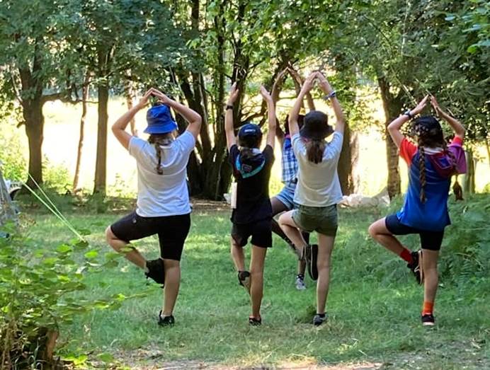 5 Guides do the tree yoga pose with their backs to the camera, surrounded by grass and trees