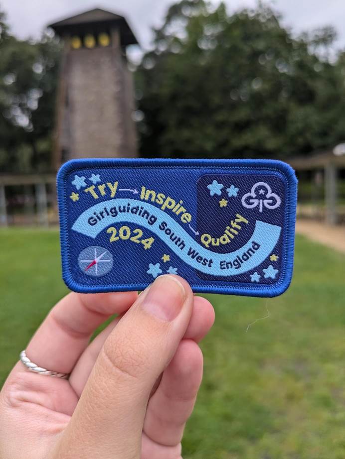 A close up of a woman's hand holding up the Try, Inspire, Qualify 2024 South West England weekend badge, which is rectangular, with a navy blue base, yellow and light blue title writing and includes a compass and a Girlguiding logo