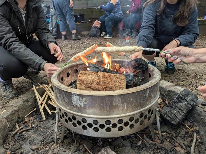 Leaders sit around a fire in the forest, holding sticks covered in dough over it, as though roasting marshmellows