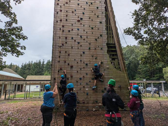 2 leaders in helmets and harnessing are on a climbing wall outside. 5 leaders on the ground, also in helmets and with harnesses, watch them and hold their safety ropes.