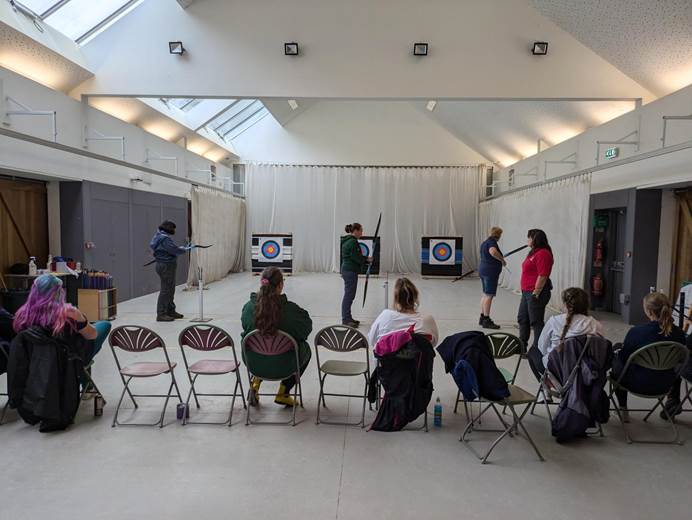 In a hall three leaders stand in front of three bullseyes ready to shoot their bows. An instructor stands to the left of them, with other leaders behind them in chairs watching.