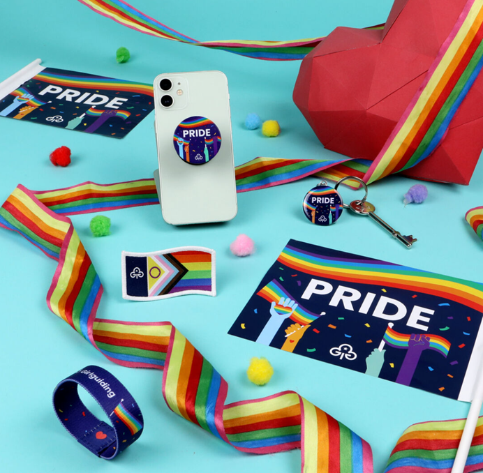 Products from Girlguiding's Pride range, including flags, a keyring, a badge and a phone grip. They all have designs with hands holding up rainbow flags with the word Pride and the Girlguiding trefoil.