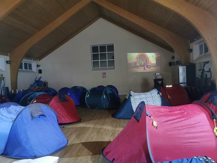 About 10 small tents are in a church hall. There is a wall behind them. On the wall, a movie is being projected