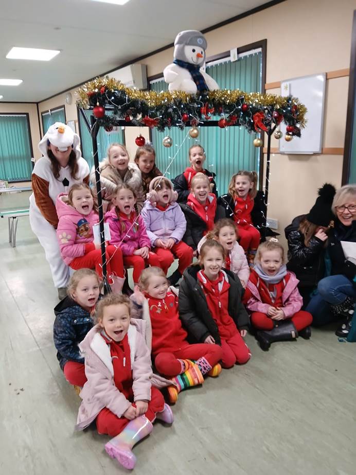 A group of Rainbows sit in a hall indoors, singing and looking at the camera smiling. A leader behind them wears a snowman suit.