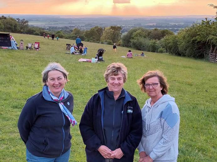 Three women stand at the front of the photo, smiling at the camera. Behind them is a field with lots of people on, then trees and more fields. The sky is orange