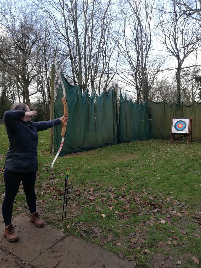 Victoria stands drawing a bow and taking aim at archery target