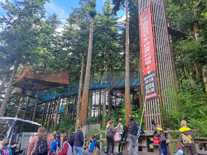 Girls and leaders arrive at Zip World in Wales. Above them is a big title sign and walkways in trees.