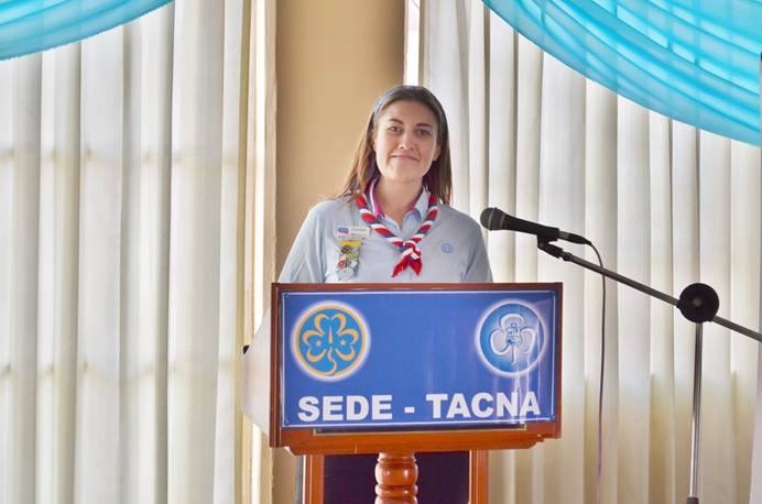 A woman with brown hair wearing a leader uniform shirt, and a red, white and blue necker, stands at a lectern smiling at the camera