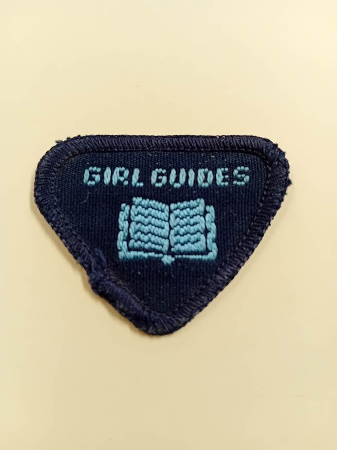 Badge in the shape of an upside triangle with the words Girl Guides and an open book embroided on the front.