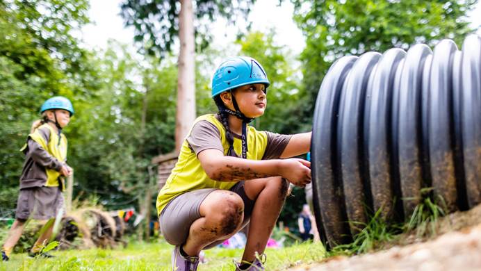 A Brownie wearing a helmet is kneeling on the ground in a field. She's about to go into a plastic tunnel.