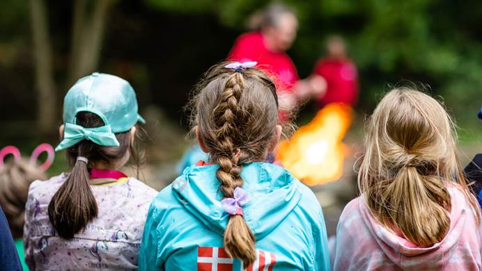 Three girls with their backs to the camera are facing towards a campfire