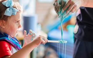 A girl in Rainbow uniform is standing in front of a bowl at a table. Her hands are covered in blue goo. There is a leader standing next to her, whose torso can be seen in the image. Her hands are also covered in blue goo.