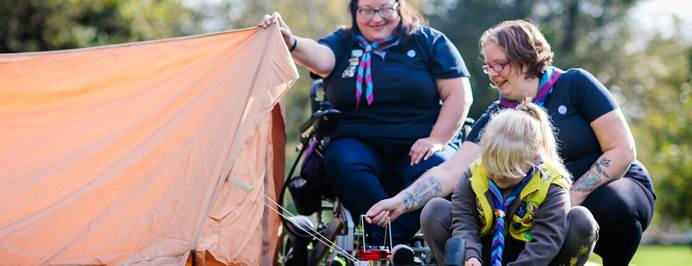 Two volunteers and one girl in Brownie uniform are assembling a tent outdoors